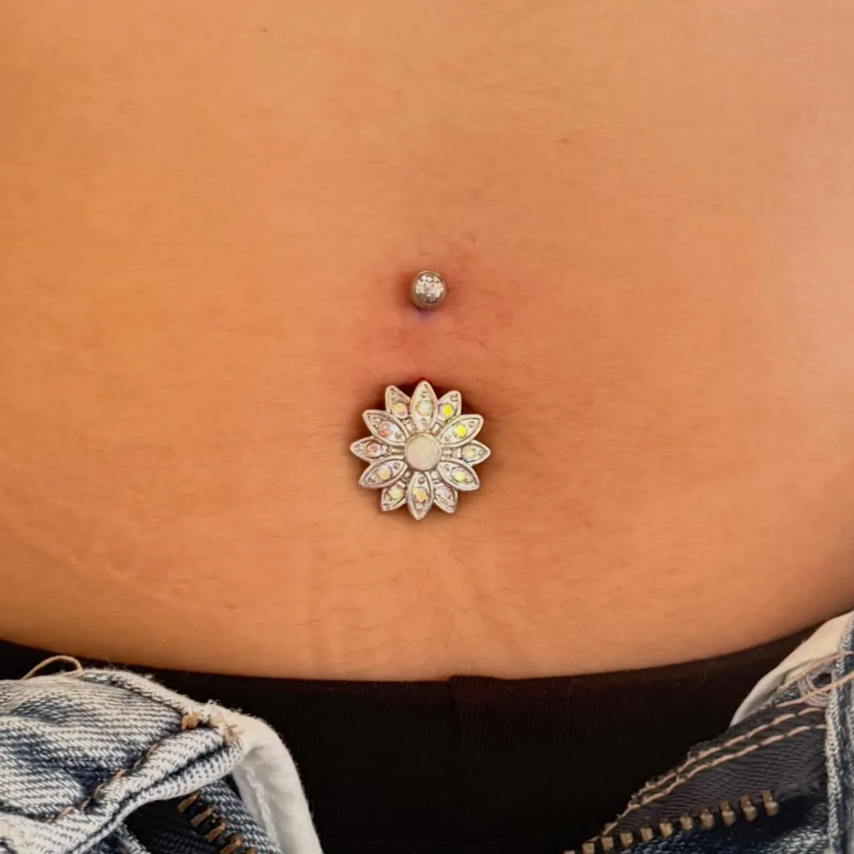 Navel piercing with a silver flower-shaped charm, opal center and iridescent petals